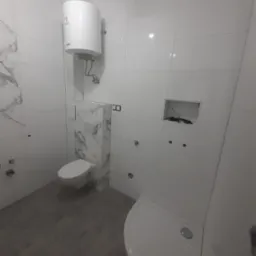 Nowa, jasna łazienka z białymi płytkami imitującymi marmur, podwieszaną toaletą, białą kabiną prysznicową i bojlerem. Widoczne przygotowane otwory na armaturę.