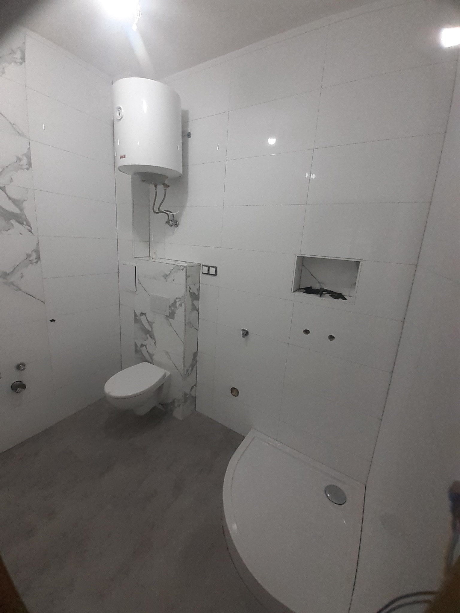 Nowa, jasna łazienka z białymi płytkami imitującymi marmur, podwieszaną toaletą, białą kabiną prysznicową i bojlerem. Widoczne przygotowane otwory na armaturę.