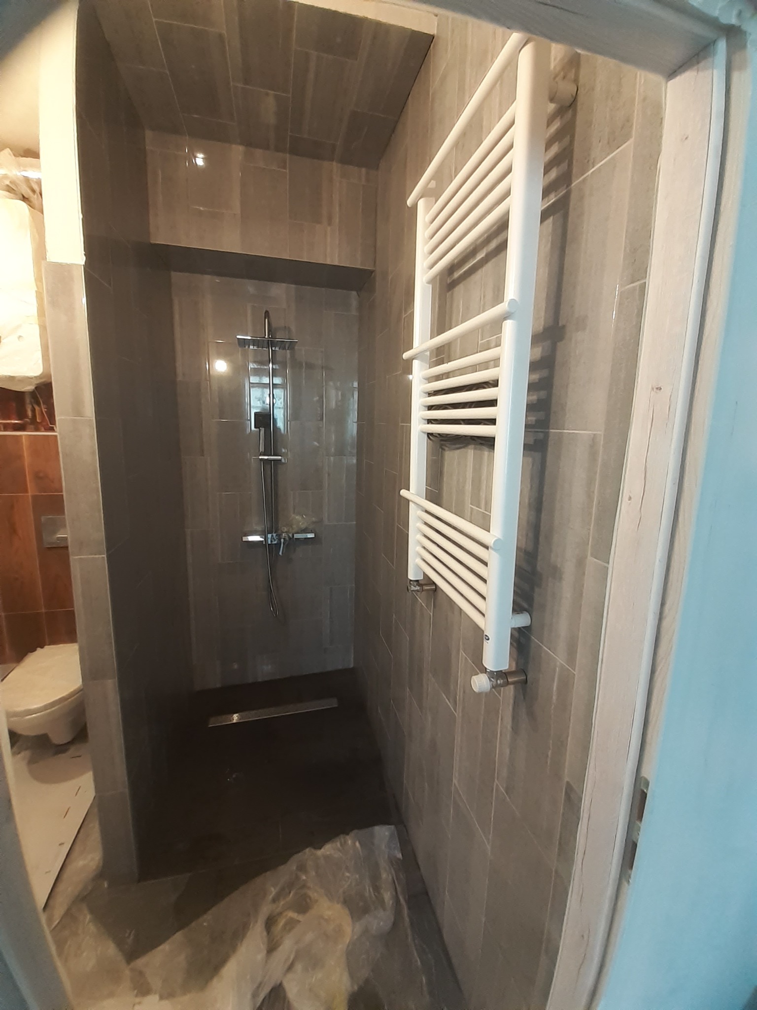 Szara kabina prysznicowa z odpływem liniowym i białym grzejnikiem drabinkowym w Ząbkowicach Śląskich, widok z perspektywy wejścia, podłoga częściowo zakryta folią ochronną.