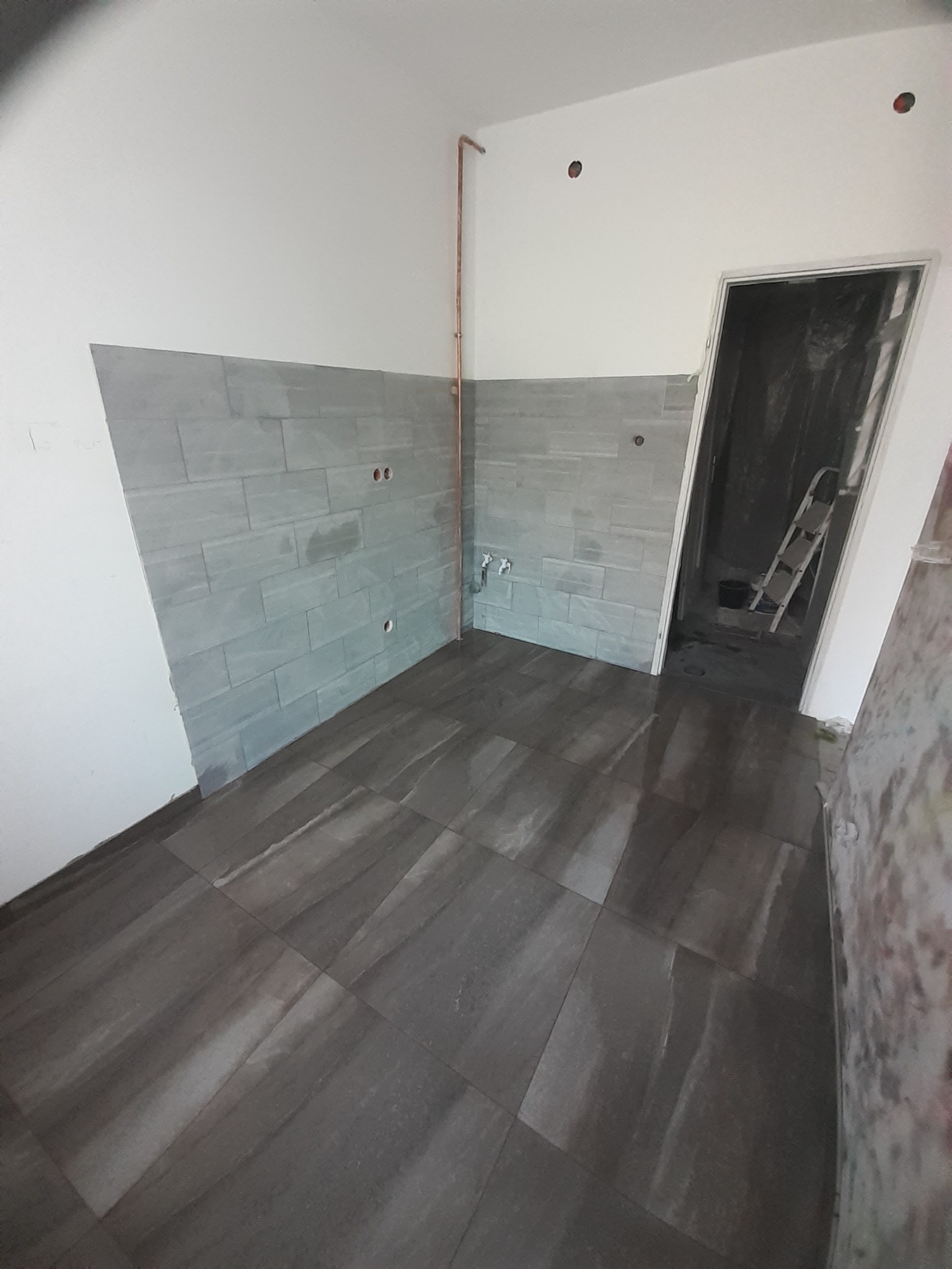 Wnętrze w trakcie remontu: ułożone ciemne płytki podłogowe, szare płytki ścienne w cegiełkę, widoczne instalacje wodne i otwory w ścianach, drabina w głębi pomieszczenia.
