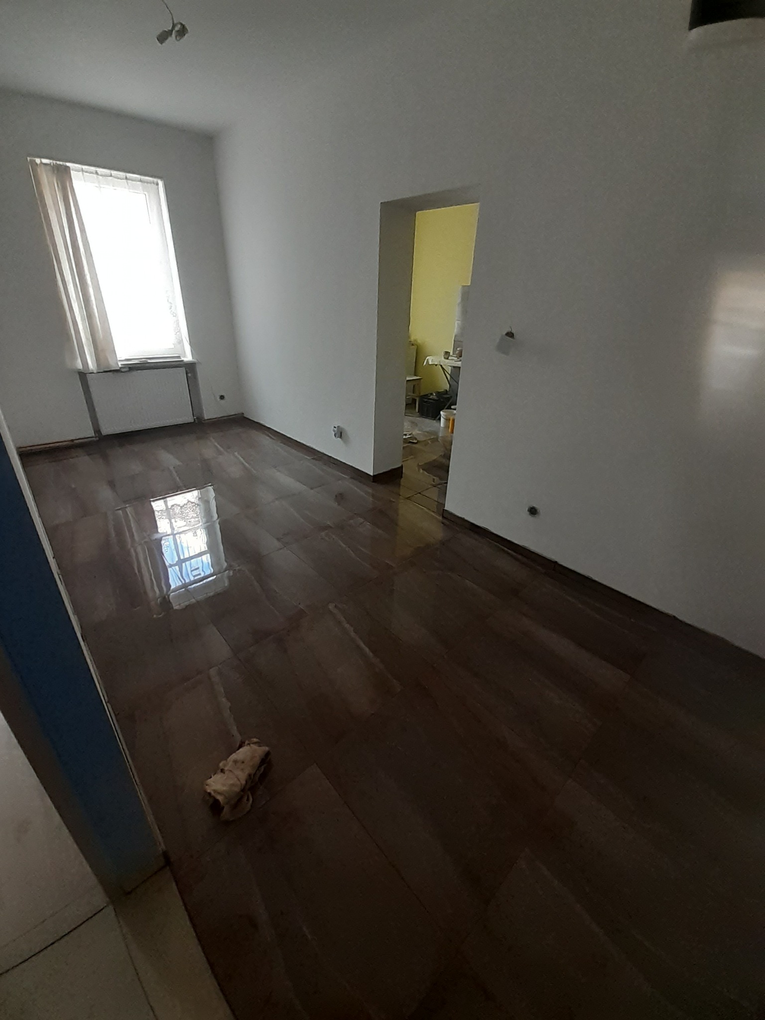 Świeżo ułożona, lśniąca, ciemnobrązowa podłoga z płytek imitujących drewno w pustym pokoju z oknem i grzejnikiem; szmatka do czyszczenia leży na podłodze.