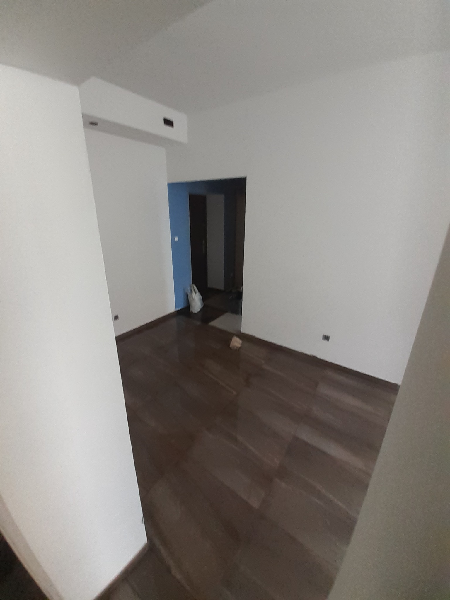 Pusta przestrzeń wewnętrzna z brązową podłogą imitującą drewno, białymi ścianami i otwartym przejściem do pokoju z niebieską ścianą. Widoczny worek foliowy w rogu pokoju.