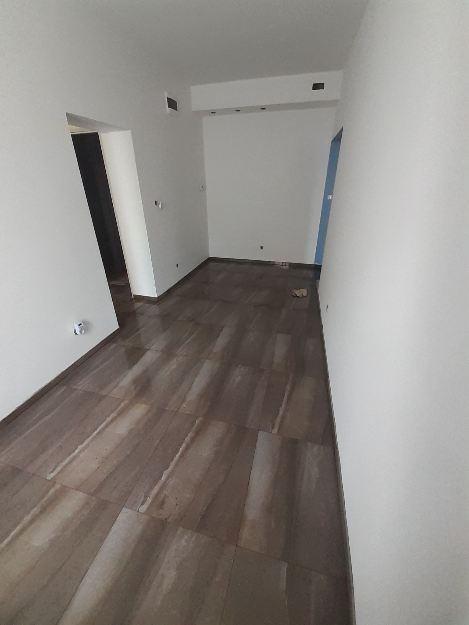 Wnętrze pustego pomieszczenia z brązową, błyszczącą podłogą imitującą kamień, białymi ścianami i widocznymi otworami drzwiowymi.