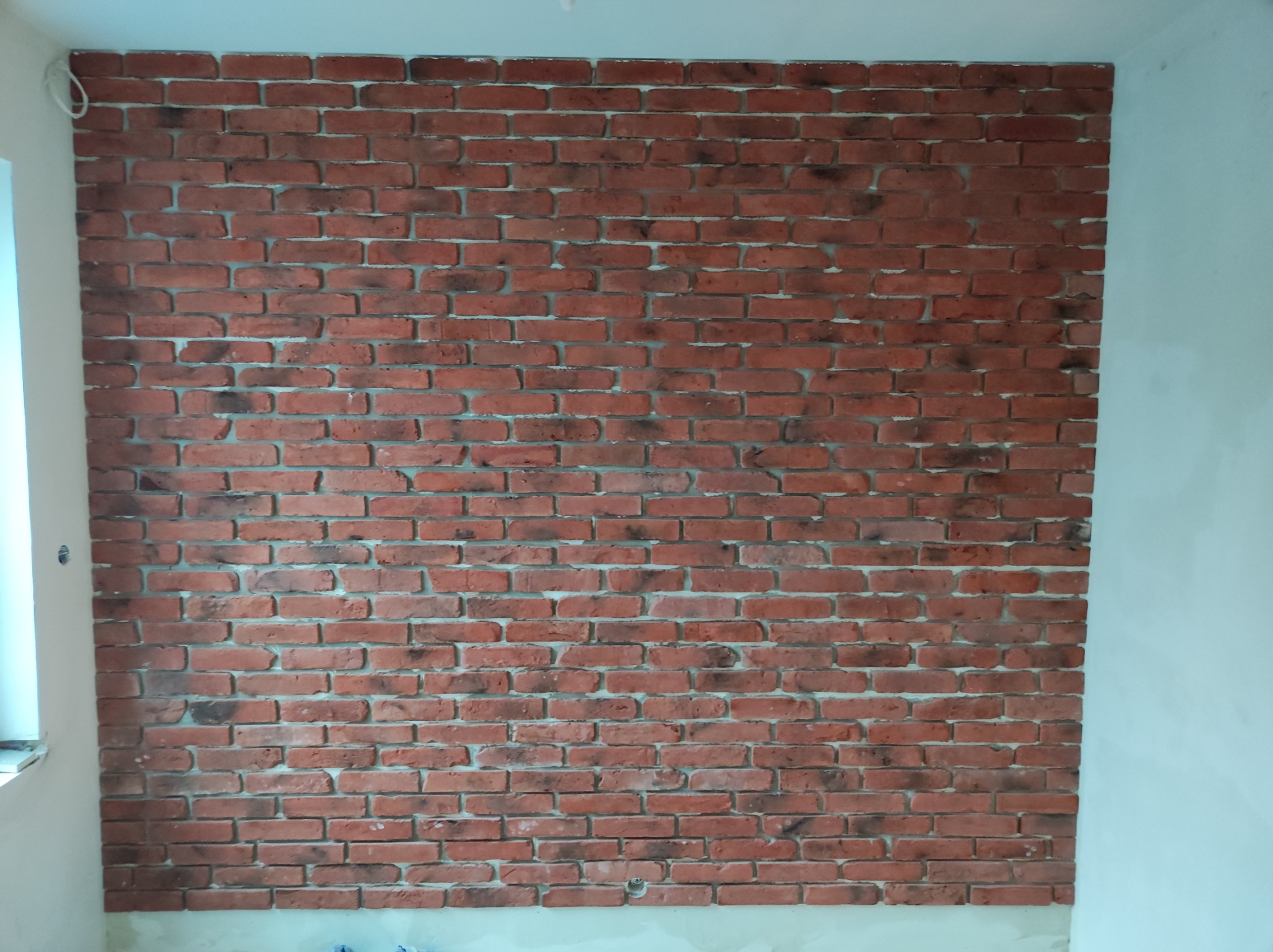 Ściana wykończona czerwoną cegłą dekoracyjną z szarymi fugami w pomieszczeniu z białymi ścianami, widoczny fragment okna po lewej stronie.