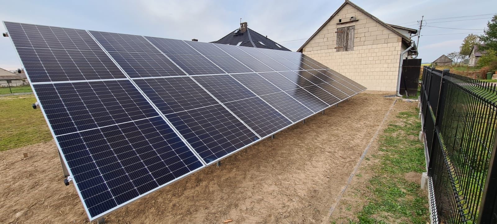 Panel słoneczny ustawiony na gruncie obok budynku gospodarczego i czarnego ogrodzenia z siatki.
