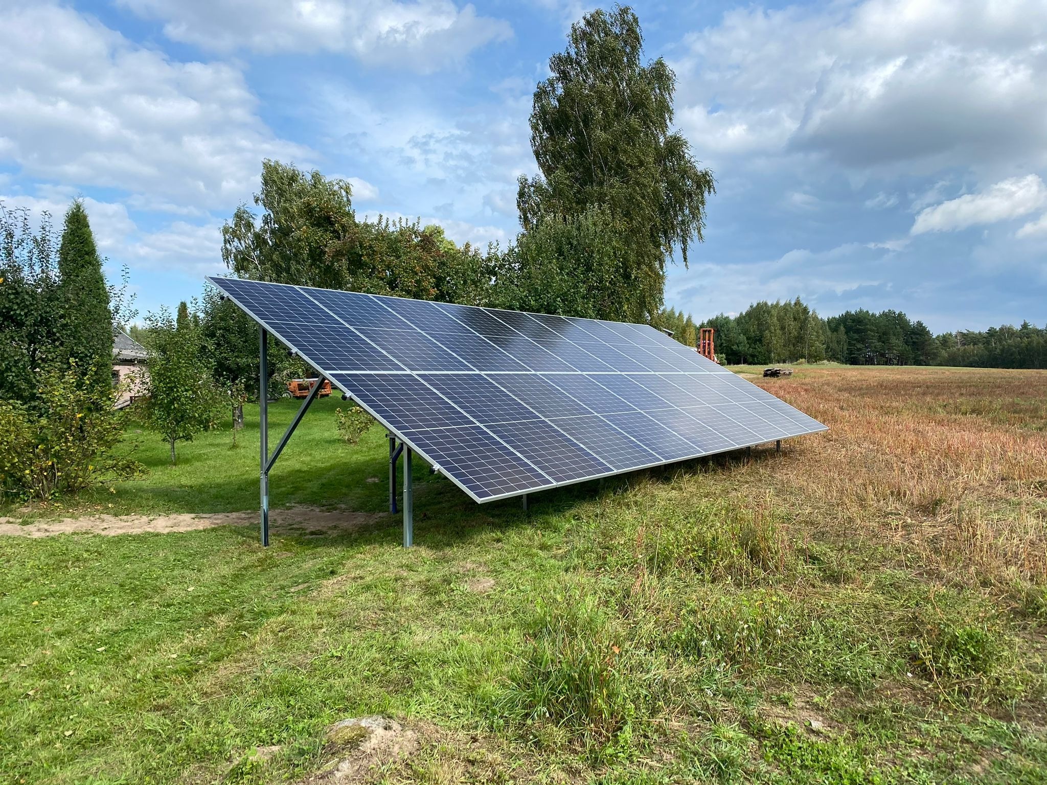 Panel słoneczny na metalowej konstrukcji, ustawiony na trawiastym terenie z drzewami w tle, pod błękitnym niebem z chmurami.