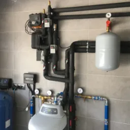 Złożona instalacja hydrauliczna z licznymi rurami w izolacji termicznej, zbiornikami wyrównawczymi i panelami sterowania, zamontowana na ścianie wyłożonej płytkami ceramicznymi.