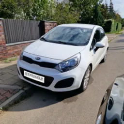 Biały samochód Kia Rio zaparkowany na ulicy w słoneczny dzień, widoczny z przodu, z fragmentem czarnego samochodu po prawej stronie kadru.