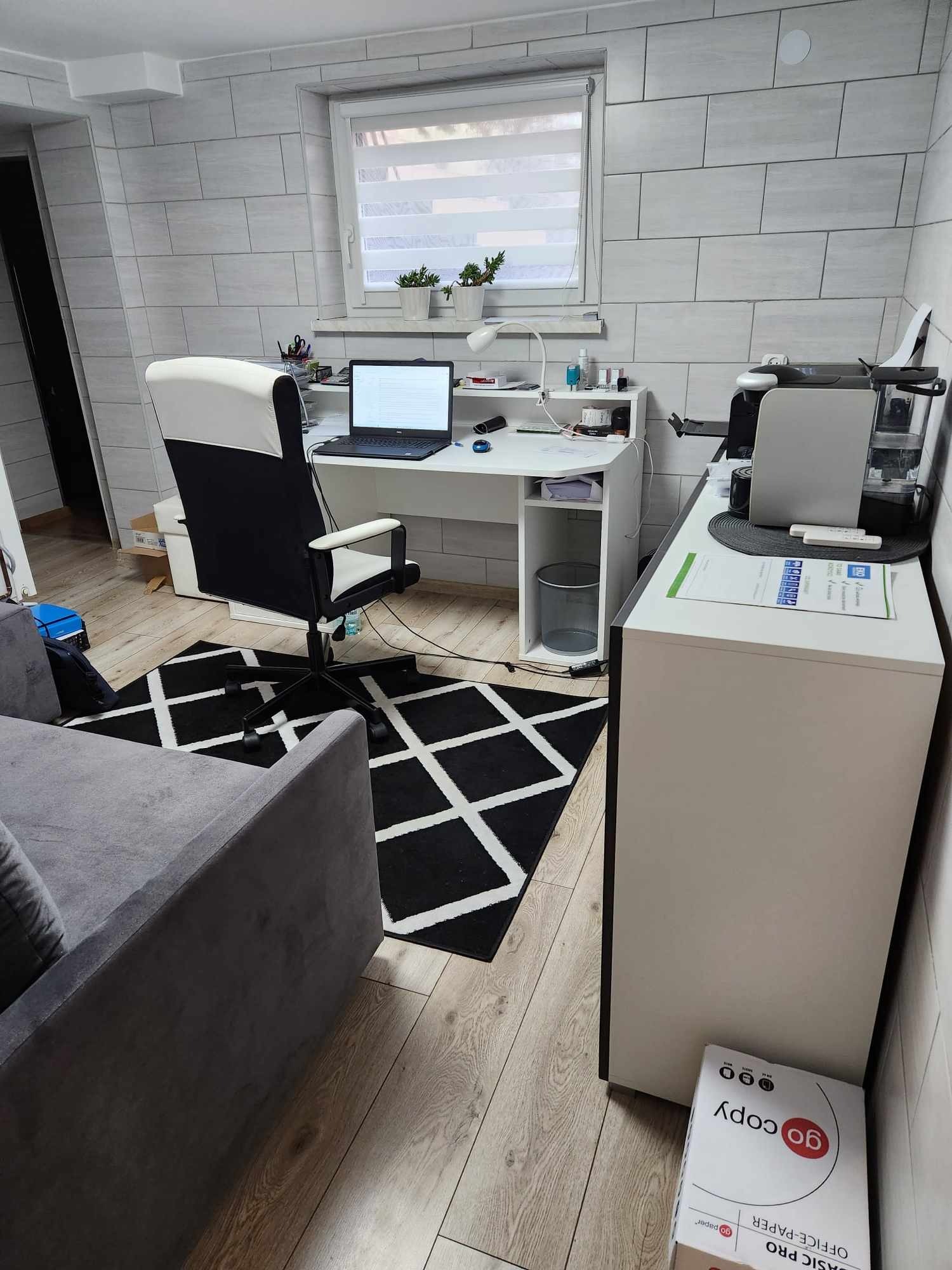 Domowe biuro z białym biurkiem, laptopem, czarno-białym krzesłem biurowym na czarno-białym dywanie w romby, z szarą sofą w rogu pokoju i białą komodą z drukarką.
