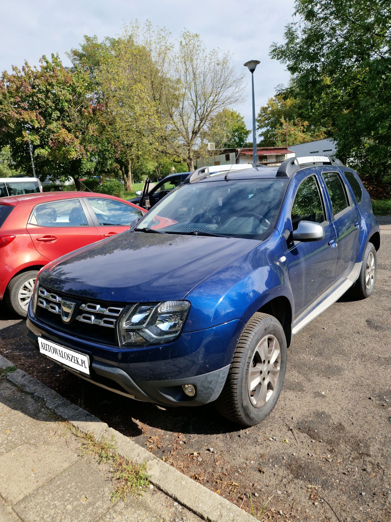 Niebieski SUV zaparkowany na placu, z widocznym logo na przedniej atrapie i tablicą rejestracyjną z adresem strony internetowej.