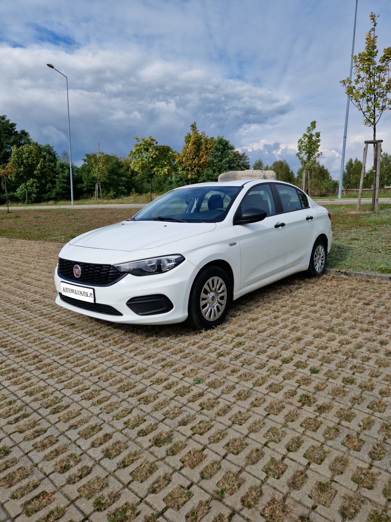 Biały samochód marki Fiat Tipo zaparkowany na ażurowym parkingu z widocznym logo firmy na tablicy rejestracyjnej i pochmurnym niebem w tle.