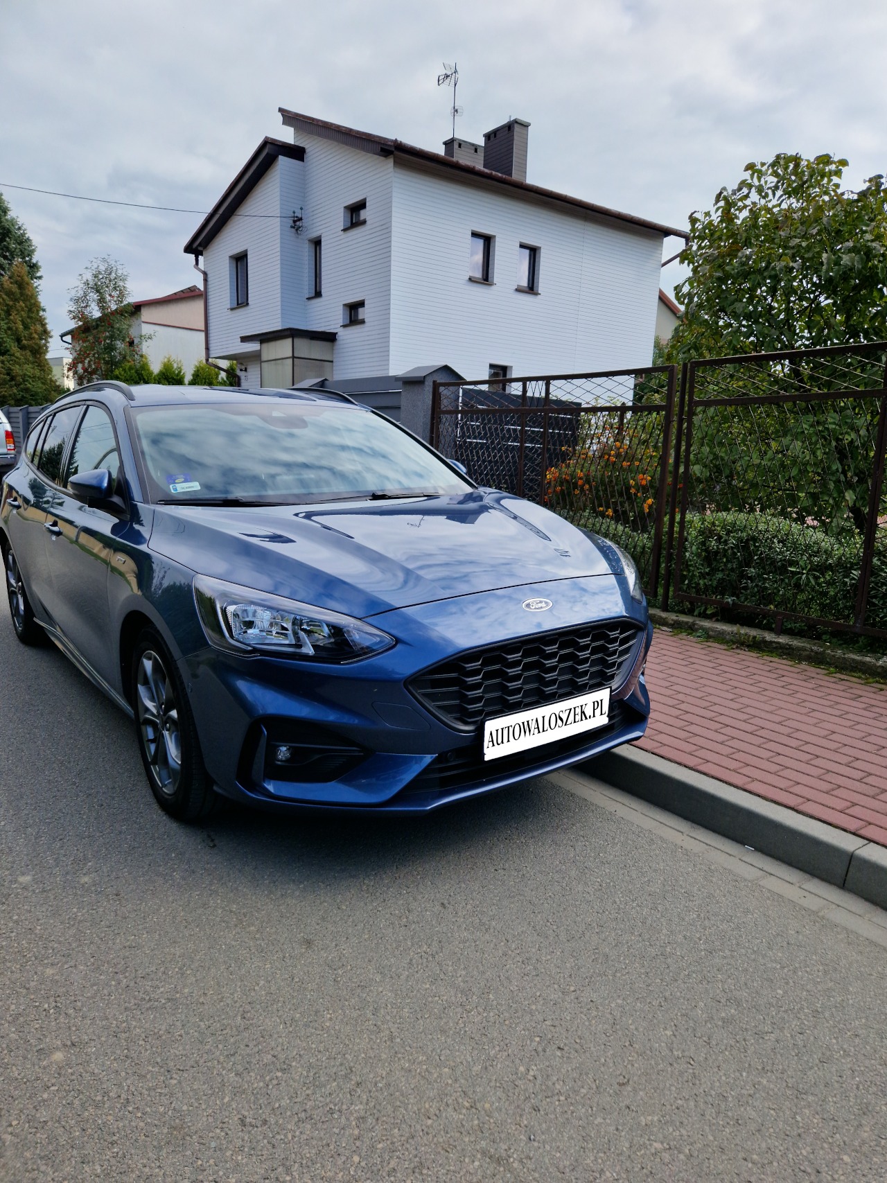 Niebieski Ford Focus kombi zaparkowany na ulicy przed białym domem z brązowymi akcentami.