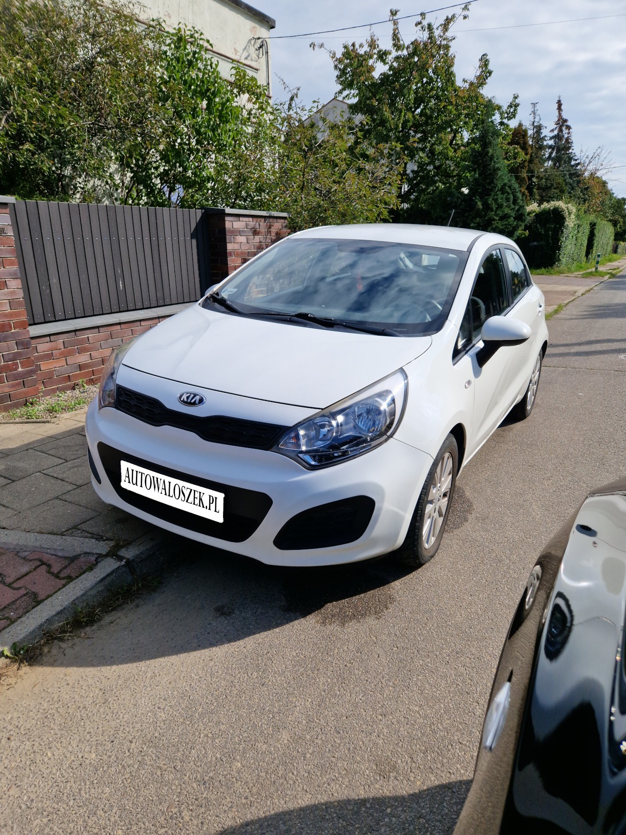 Biały samochód Kia Rio zaparkowany na ulicy w słoneczny dzień, widoczny z przodu, z fragmentem czarnego samochodu po prawej stronie kadru.