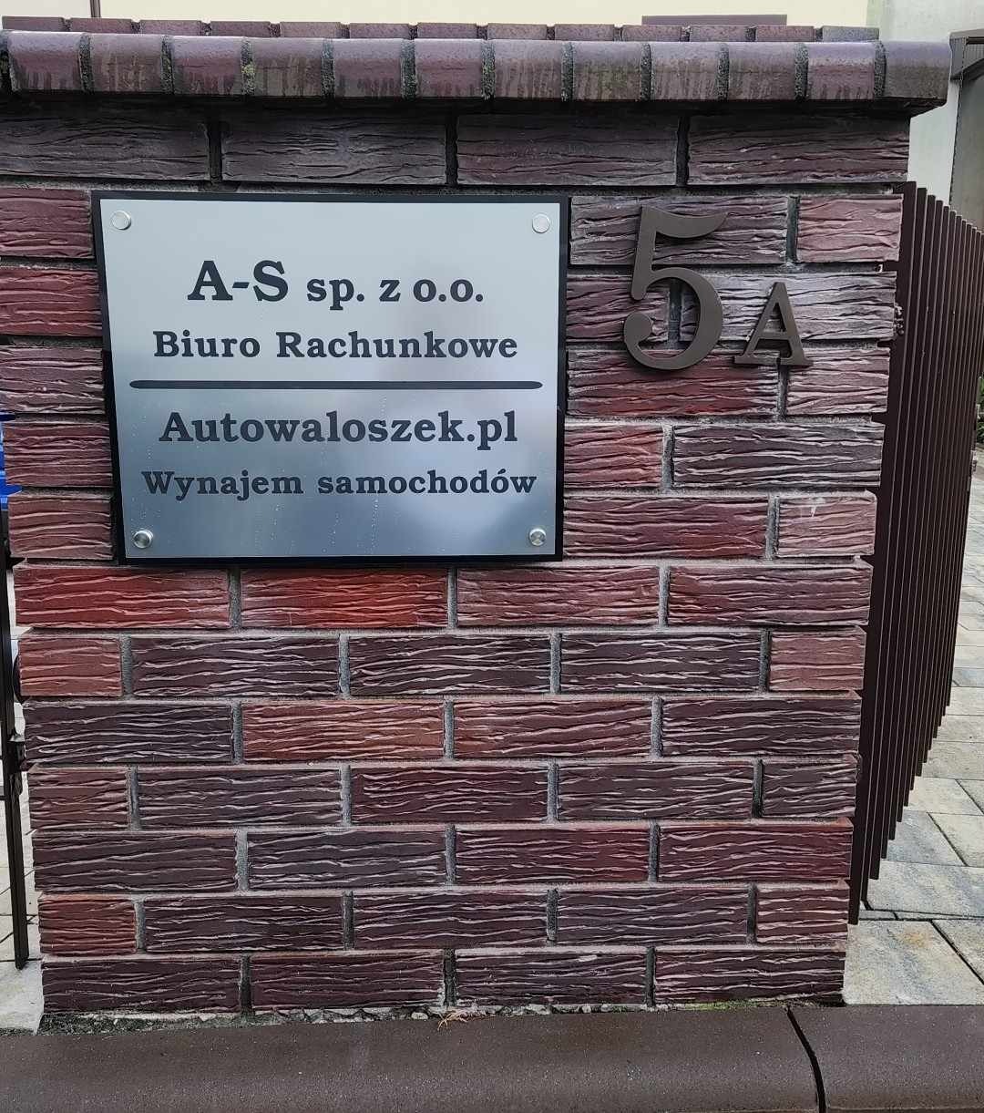 Tablica informacyjna firmy A-S sp. z o.o. Biuro Rachunkowe z adresem strony internetowej Autowaloszek.pl i dopiskiem Wynajem samochodów, umieszczona na ceglanym filarze obok numeru 5A.
