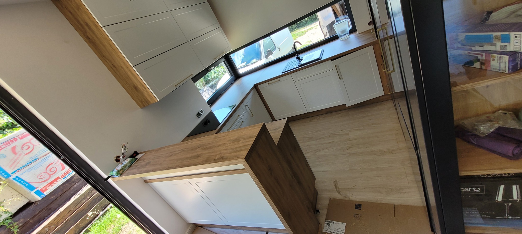 Nowoczesna kuchnia w jasnych barwach z drewnianymi akcentami na blacie i obrzeżach, widok z góry, na blacie czarny zlew i płyta indukcyjna, w tle okno z widokiem na samochód dostawczy.