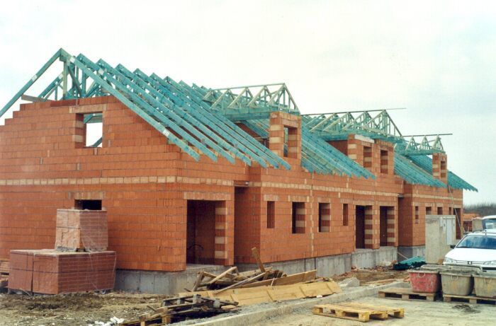 Wznoszone w stanie surowym szeregowe domy z czerwonej cegły z widoczną konstrukcją dachu z impregnowanego na niebiesko drewna, palety z cegłami i gruzem na placu budowy.