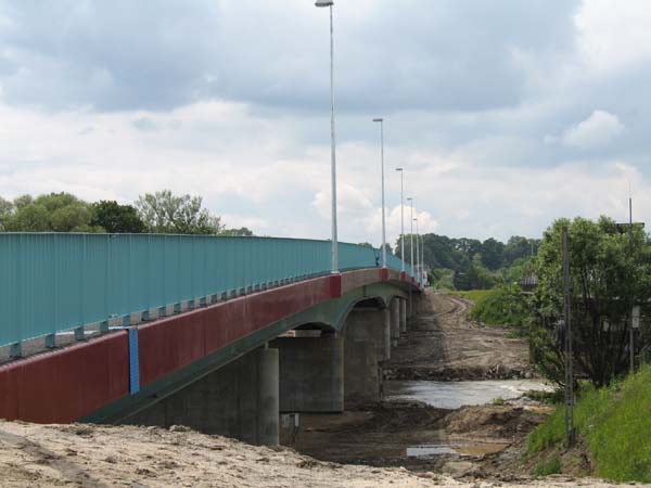 Betonowy most z niebieskimi barierkami i czerwonymi elementami konstrukcyjnymi, widoczny z dołu w trakcie budowy, z ziemią i błotem wokół filarów i drogi dojazdowej.