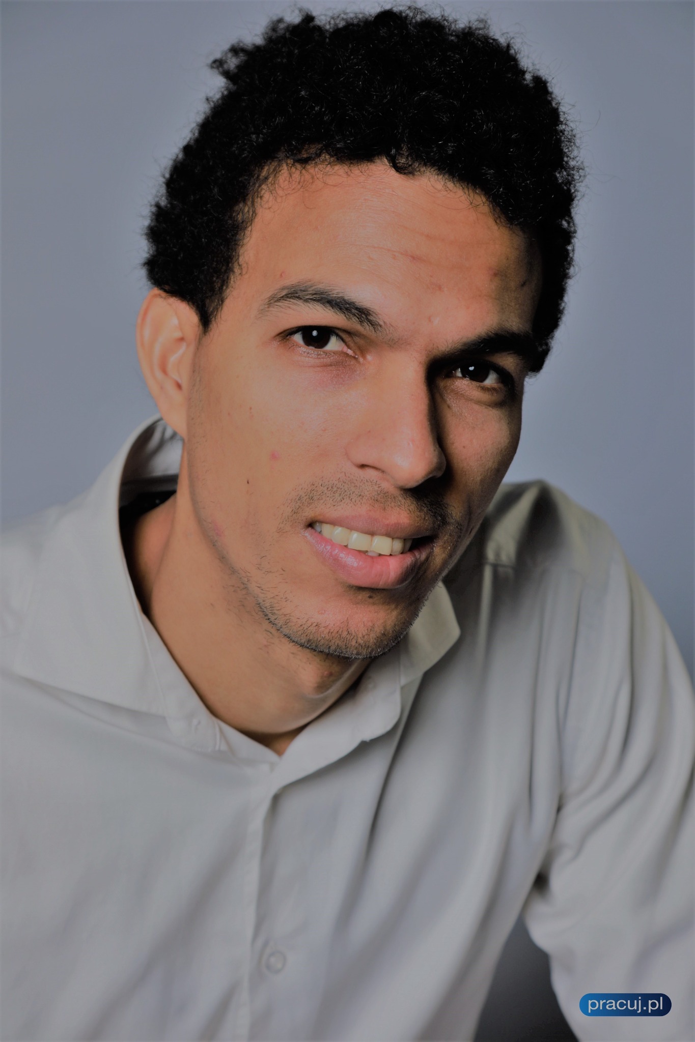 Portret uśmiechniętego młodego mężczyzny z kręconymi włosami, ubrany w jasną koszulę na tle szarego tła, z logo pracuj.pl w prawym dolnym rogu.