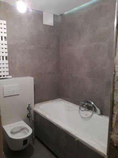 Nowoczesna łazienka z szarą płytką, białą wanną zabezpieczoną folią i podwieszaną toaletą z bidetką, oświetlenie LED podsufitowe.