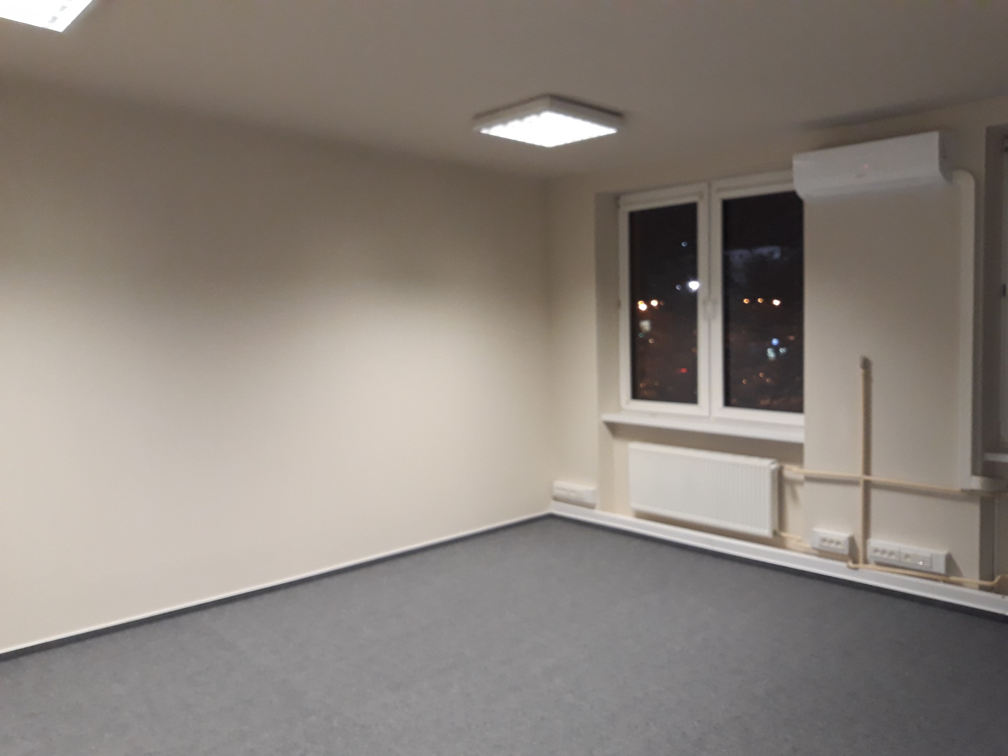 Puste biuro z szarym dywanem, białymi ścianami, grzejnikiem i klimatyzatorem pod oknem, widok na nocne miasto.