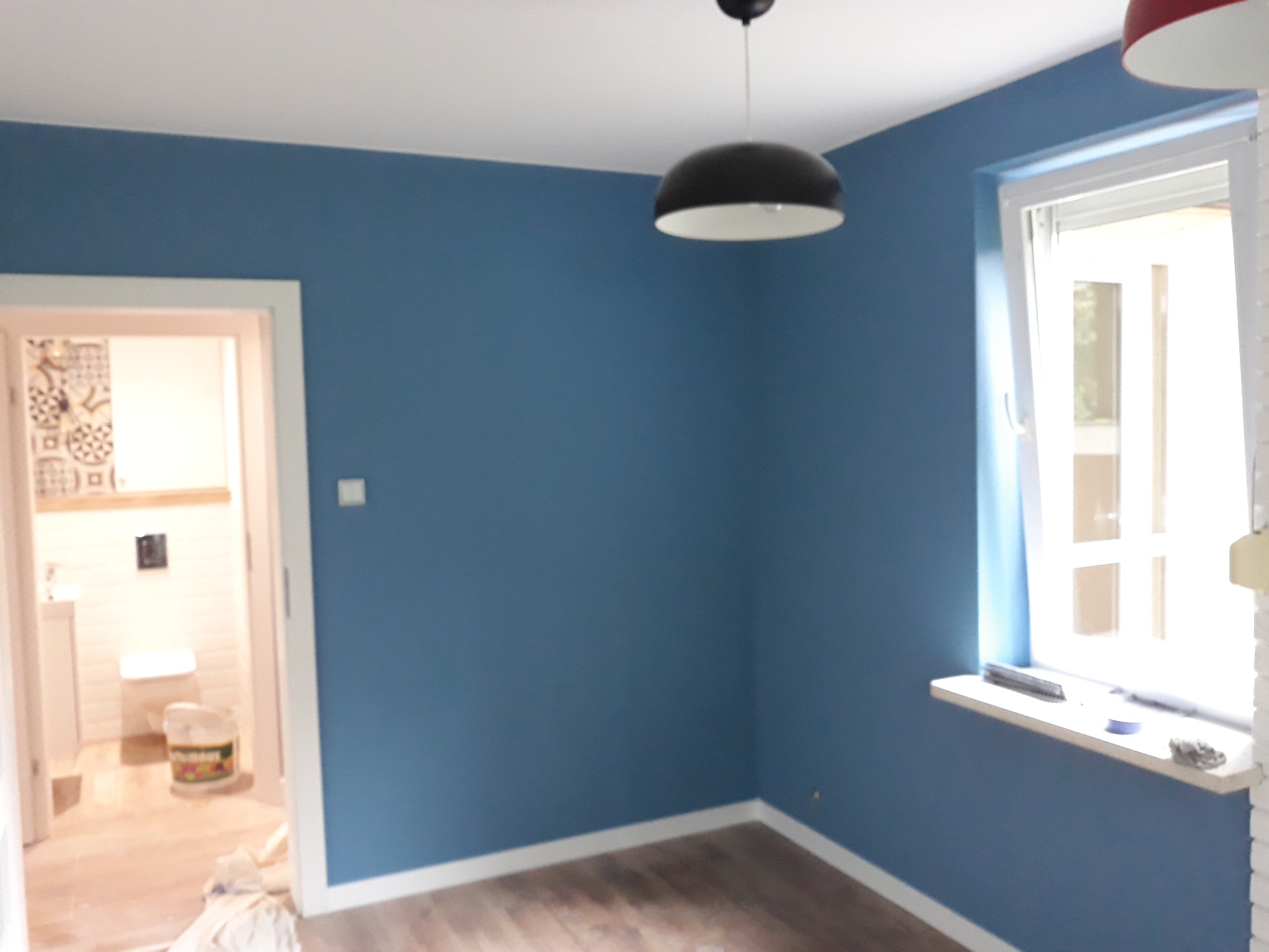 Świeżo pomalowane na niebiesko ściany w pokoju z drewnianą podłogą, widoczna łazienka w tle przez otwarte drzwi, czarna lampa sufitowa i białe okno z parapetem.