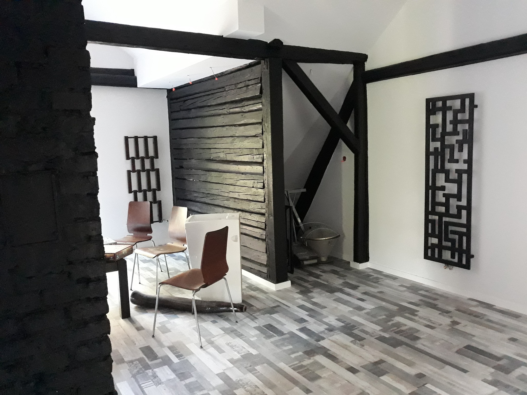 Wnętrze pokoju z drewnianą ścianką działową i czarnymi belkami stropowymi, podłoga wyłożona panelami imitującymi drewno, widoczne krzesła i dekoracyjny grzejnik.