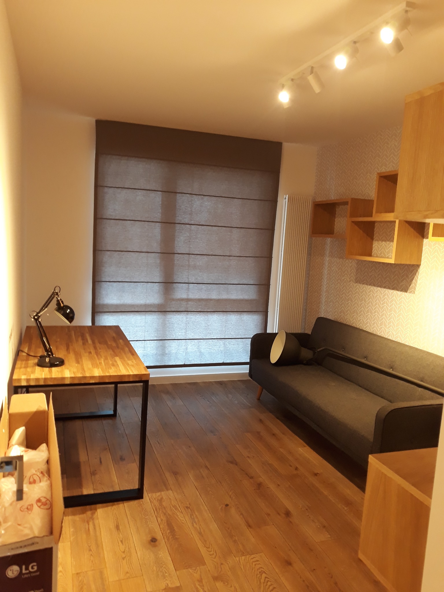 Jasne wnętrze pokoju z drewnianą podłogą, meblami w stylu skandynawskim, szarą sofą i biurkiem z lampką, roletą rzymską w oknie oraz kartonem po telewizorze LG.