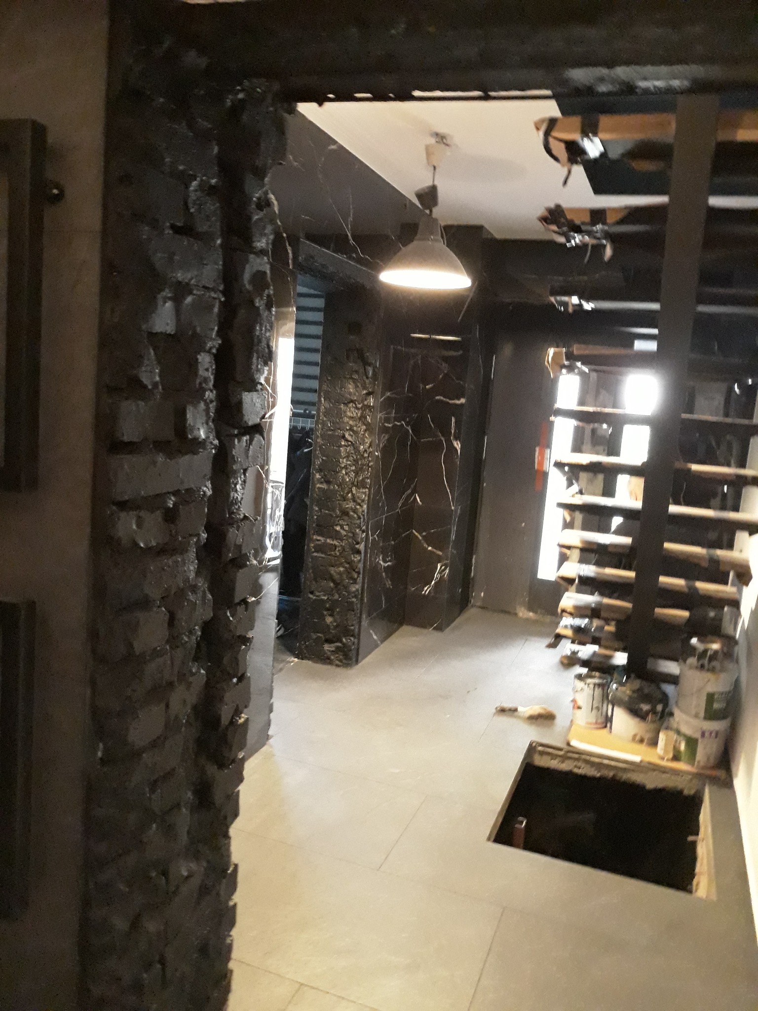 Wnętrze w trakcie prac wykończeniowych: czarna cegła dekoracyjna na ścianie, marmurowe wykończenie, schody z surowego drewna, widoczne puszki z farbą i otwór w podłodze.