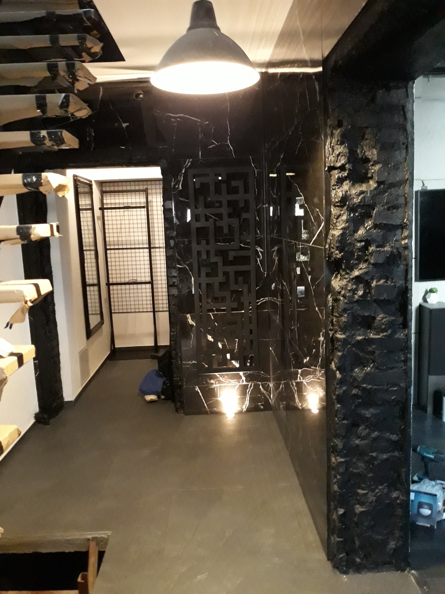 Wnętrze w trakcie prac, widoczna czarna marmurowa ściana z dekoracyjnym ażurem, sufit z elementami w trakcie montażu, fragment ściany z surowej cegły pomalowanej na czarno, podłoga w szarym...