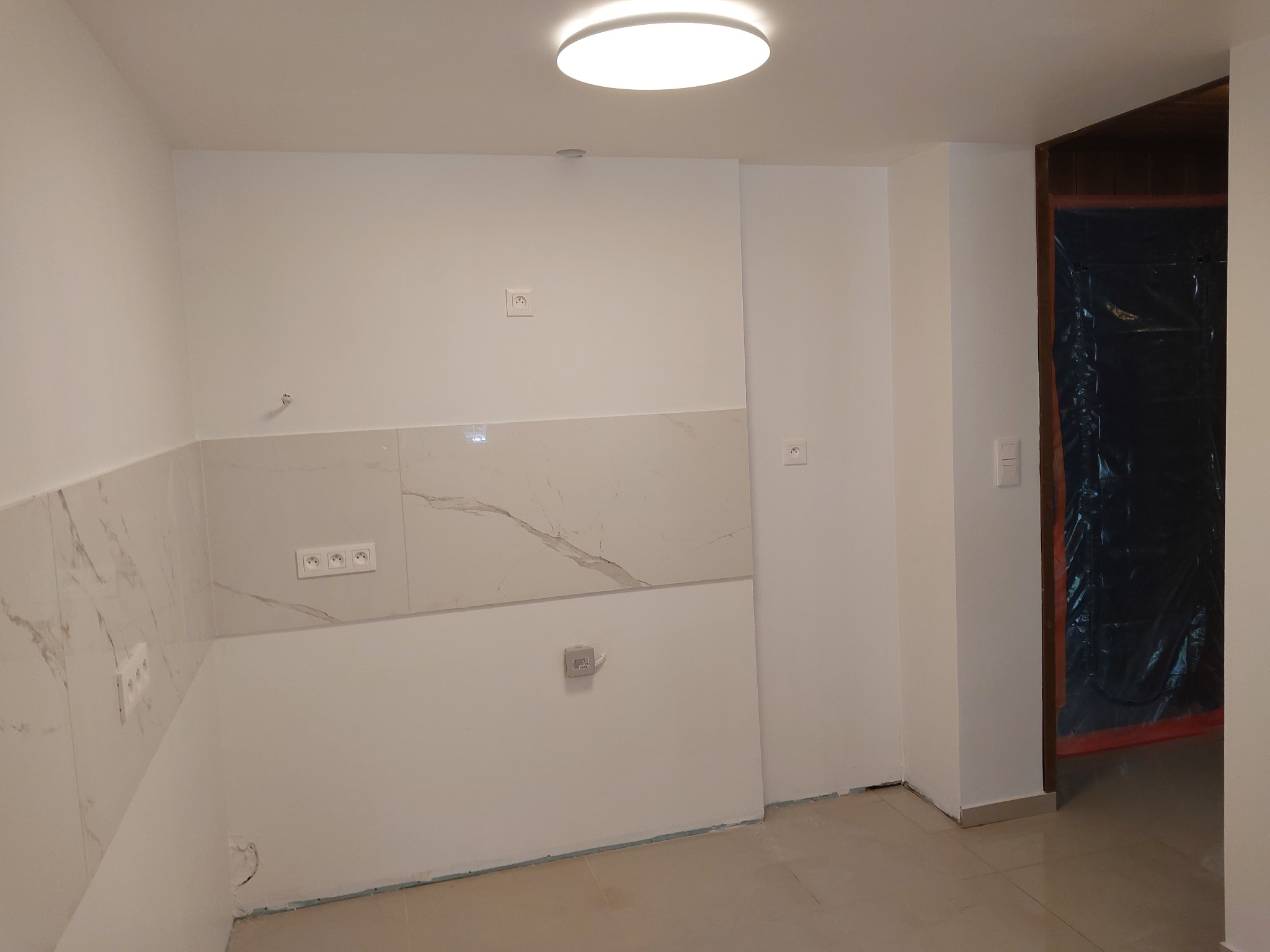 Przygotowane do użytku, jasne wnętrze z białymi ścianami, marmurowymi płytkami nad blatem, gniazdkami elektrycznymi i zabezpieczonym wejściem do innego pomieszczenia folią ochronną.