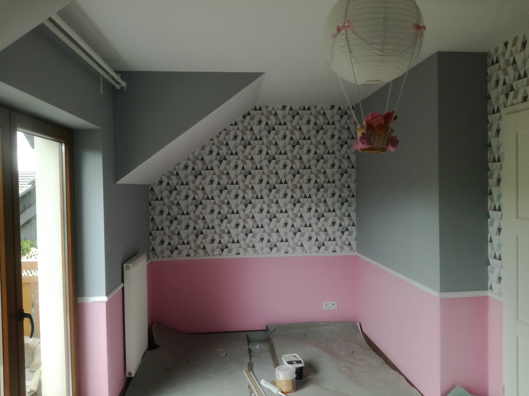 Pokój dziecięcy w trakcie odnawiania: ściany pomalowane na szaro i różowo, fragment ściany z tapetą w geometryczne wzory, na podłodze folia ochronna i puszki z farbą, pod sufitem ozdobna lampa...