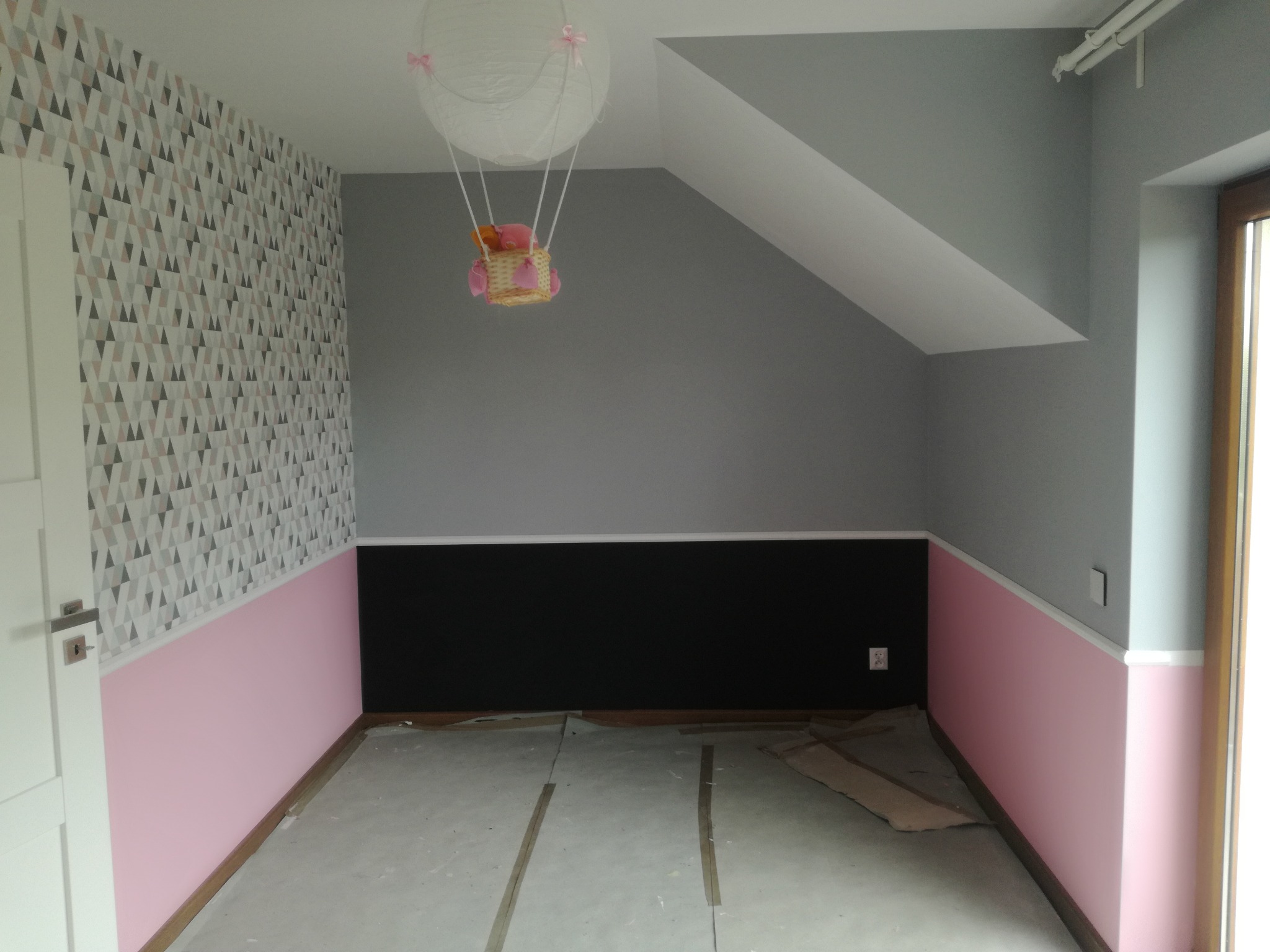 Pokój dziecięcy w trakcie remontu: ściana z geometrycznym wzorem, dolna część pomalowana na różowo, górna na szaro, czarny pas na środku, sufit z dekoracją w kształcie balonu, podłoga zabezpieczona...