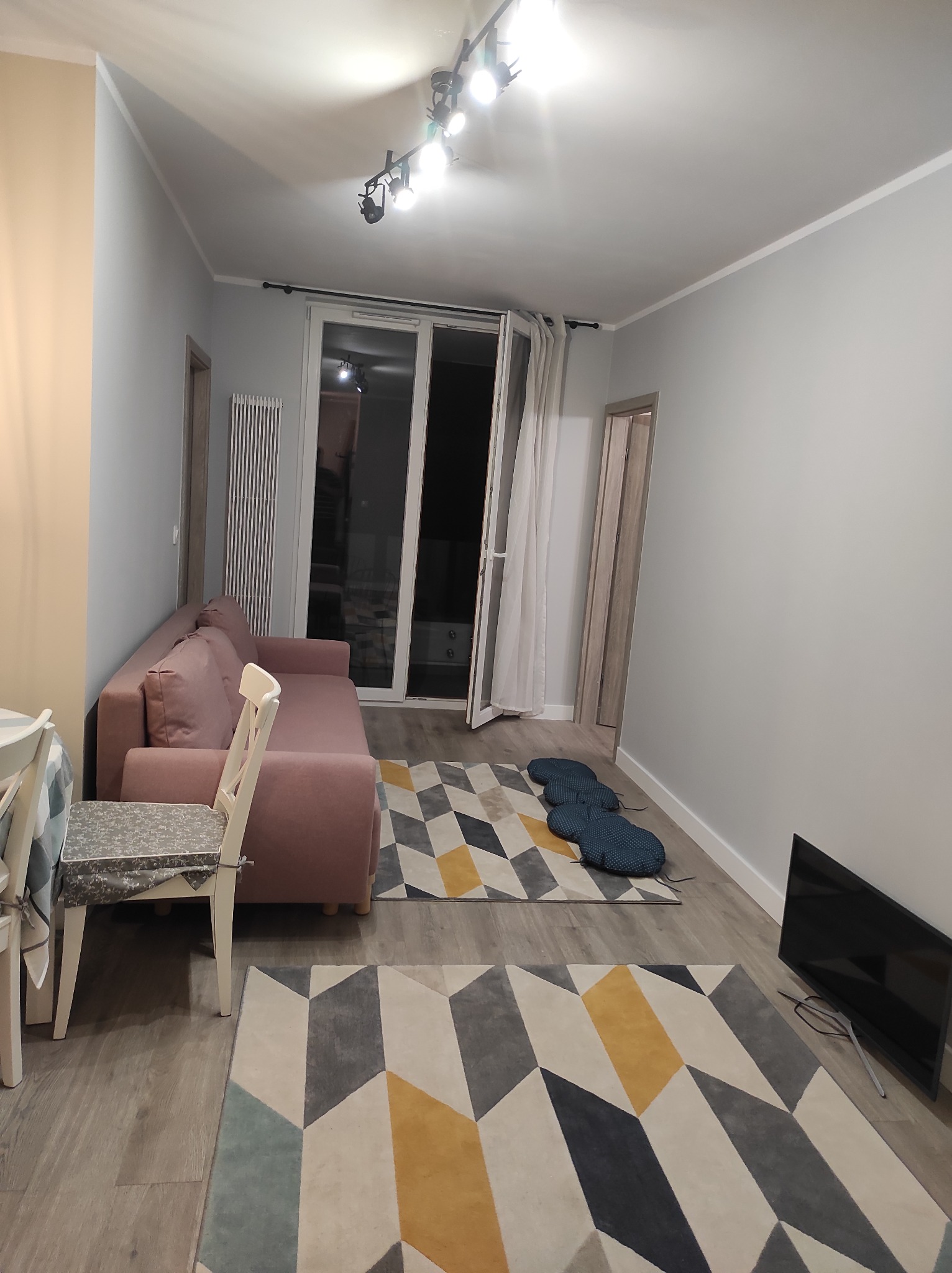 Ustawne wnętrze pokoju dziennego z szarą kolorystyką ścian, podłogi i dywanu w geometryczne wzory, z różową sofą i białymi krzesłami przy stole, oświetlone lampami sufitowymi.