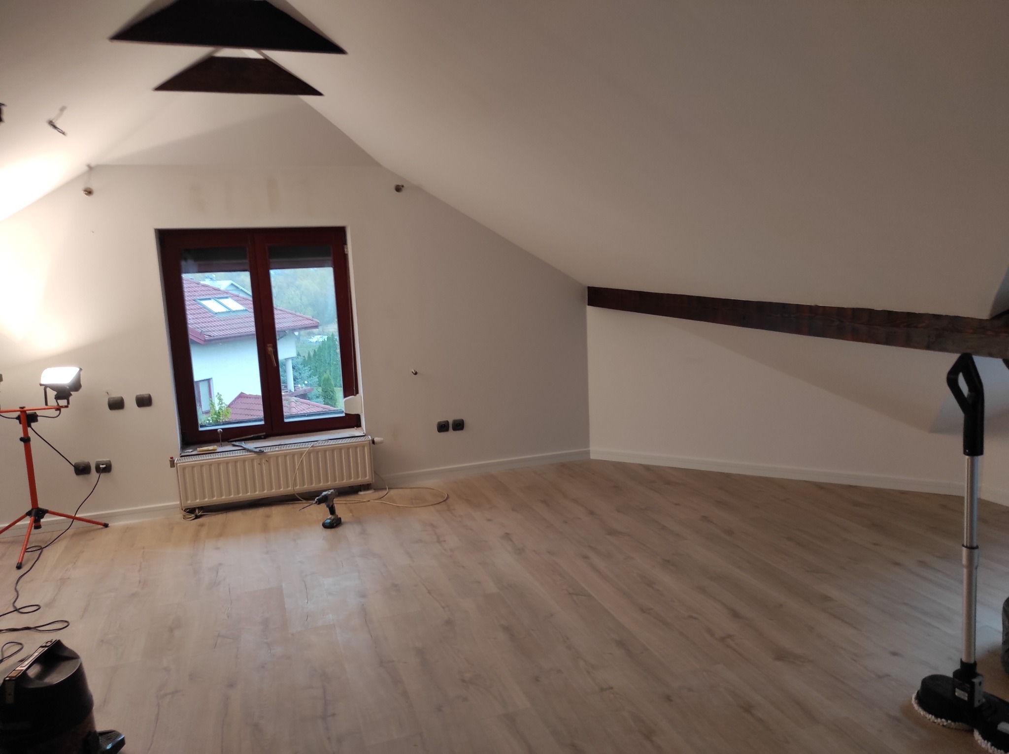 Poddasze po położeniu paneli podłogowych, widoczny fragment sufitu z drewnianymi elementami, okno, grzejnik, lampa na statywie i wiertarka akumulatorowa na podłodze.