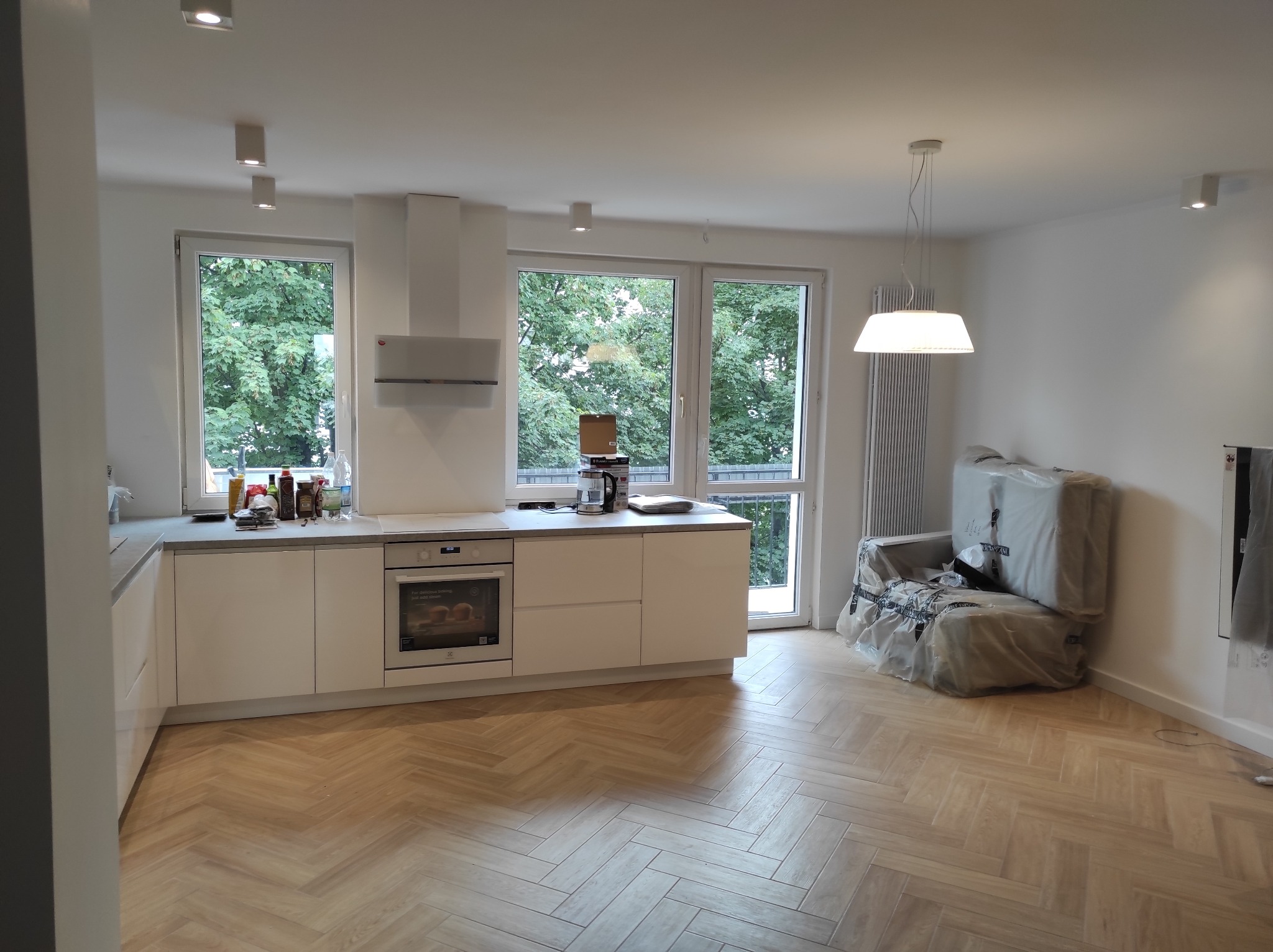 Jasna kuchnia z białymi szafkami i drewnianą podłogą w jodełkę, widok na zielone drzewa za oknem, meble zabezpieczone folią w tle.