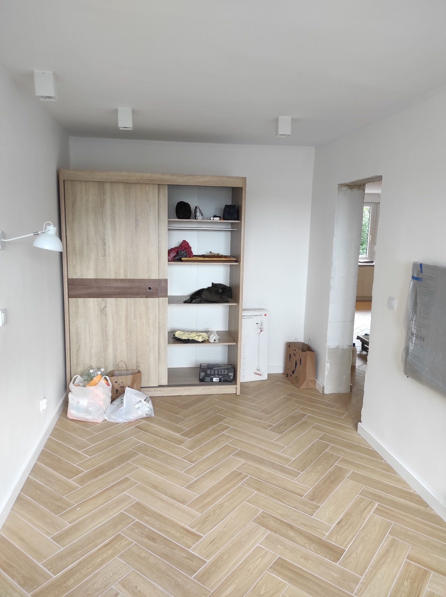 Jasny pokój z nową podłogą ułożoną w jodełkę, drewniana szafa przesuwna z częściowo otwartymi drzwiami, widoczne torby z zakupami i karton odkurzacza, ściany świeżo pomalowane na biało.
