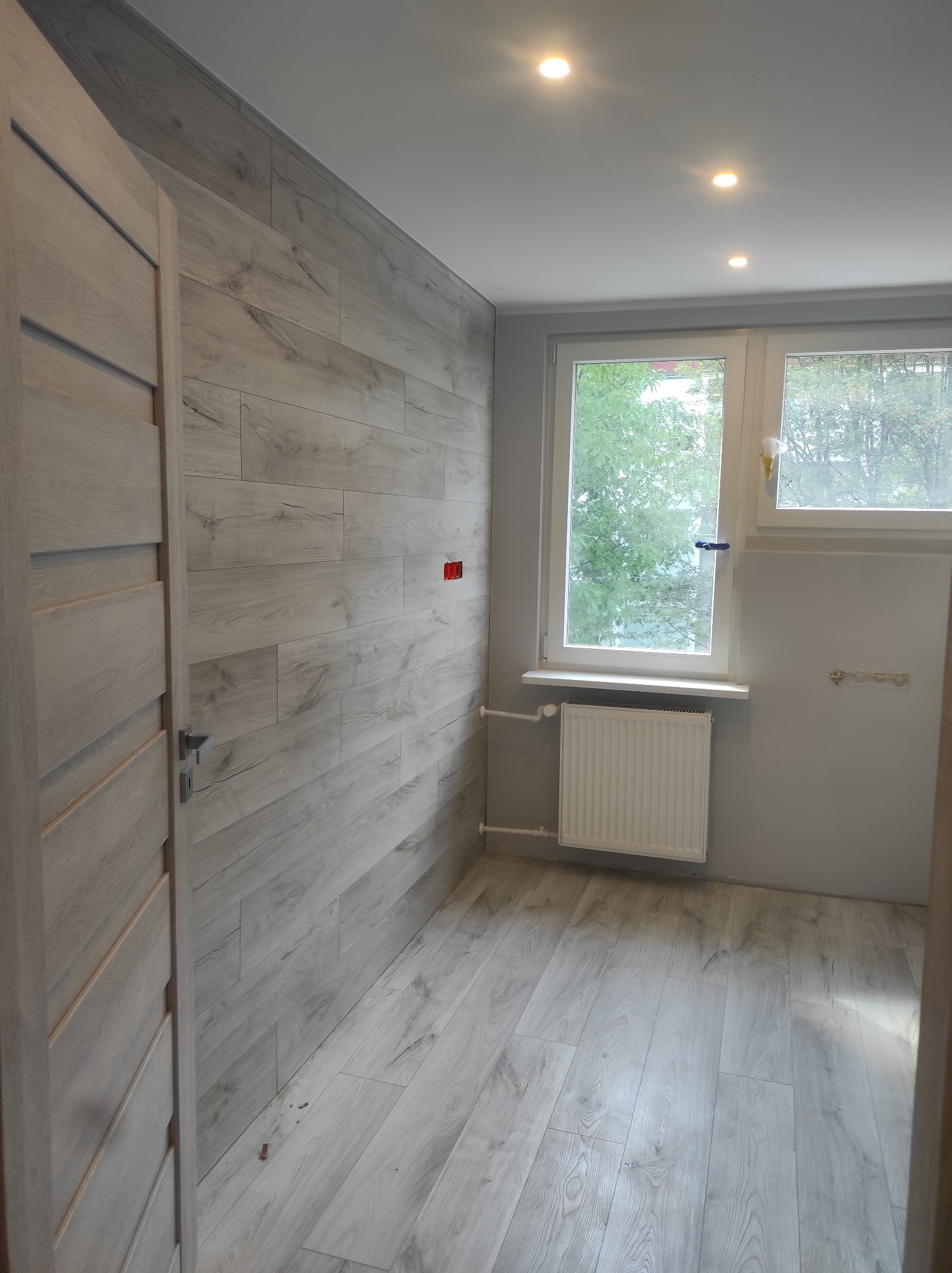 Wnętrze pokoju z panelami ściennymi imitującymi drewno, widoczne okno z grzejnikiem, zamontowane drzwi wewnętrzne oraz punkty oświetleniowe w suficie.