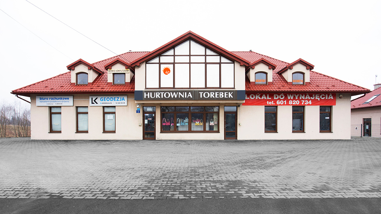 Budynek parterowy z czerwonym dachem, na którym znajdują się szyldy: Biuro Rachunkowe, Geodezja, Hurtownia Torebek, Lokal do wynajęcia. Na wystawie Hurtowni Torebek widoczne torebki.