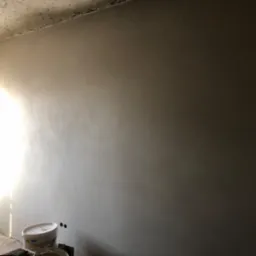 Świeżo położona, szara ściana w trakcie prac wykończeniowych, widoczna drabina i wiadro po gładzi, w rogu ściany widoczne otwory na gniazdka elektryczne.