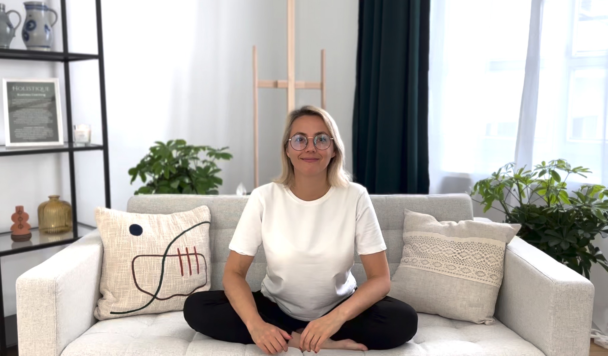 Kobieta w okularach siedzi w pozycji lotosu na kanapie w jasnym pokoju, za nią regał i sztaluga.