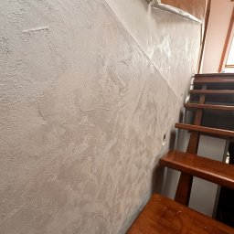 REMONT Z PASJĄ Patryk Kik - Dekoracyjna ściana z efektem stiuku w odcieniach szarości, sąsiadująca z drewnianymi schodami. Widoczne zabezpieczenie folią malarską.