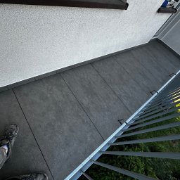 REMONT Z PASJĄ Patryk Kik - Szare płytki na balkonie ułożone równolegle do ściany, widok z góry, fragment balustrady i butów osoby wykonującej pracę. Widoczne połączenia między płytkami.