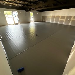 REMONT Z PASJĄ Patryk Kik - Sala z szarymi matami podłogowymi, przygotowana do ćwiczeń. Ściany częściowo wykończone, sufit w ciemnym kolorze. Widoczne okna z jasnym światłem dziennym.
