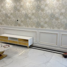 REMONT Z PASJĄ Patryk Kik - Eleganckie wnętrze: białe meble z złotymi akcentami na tle wzorzystej tapety. Marmurowa podłoga i stolik kawowy dopełniają luksusowy charakter pomieszczenia.