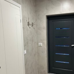 REMONT Z PASJĄ Patryk Kik - Nowoczesny przedpokój z szarą, teksturowaną płytką na ścianach, białą szafą i ciemnoszarymi drzwiami z poziomymi przeszkleniami. W tle widoczny panel sterowania.