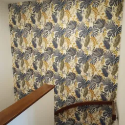 Ściana w klatce schodowej pokryta tapetą w tropikalne liście monstery i palmy, widoczna drewniana poręcz schodów.