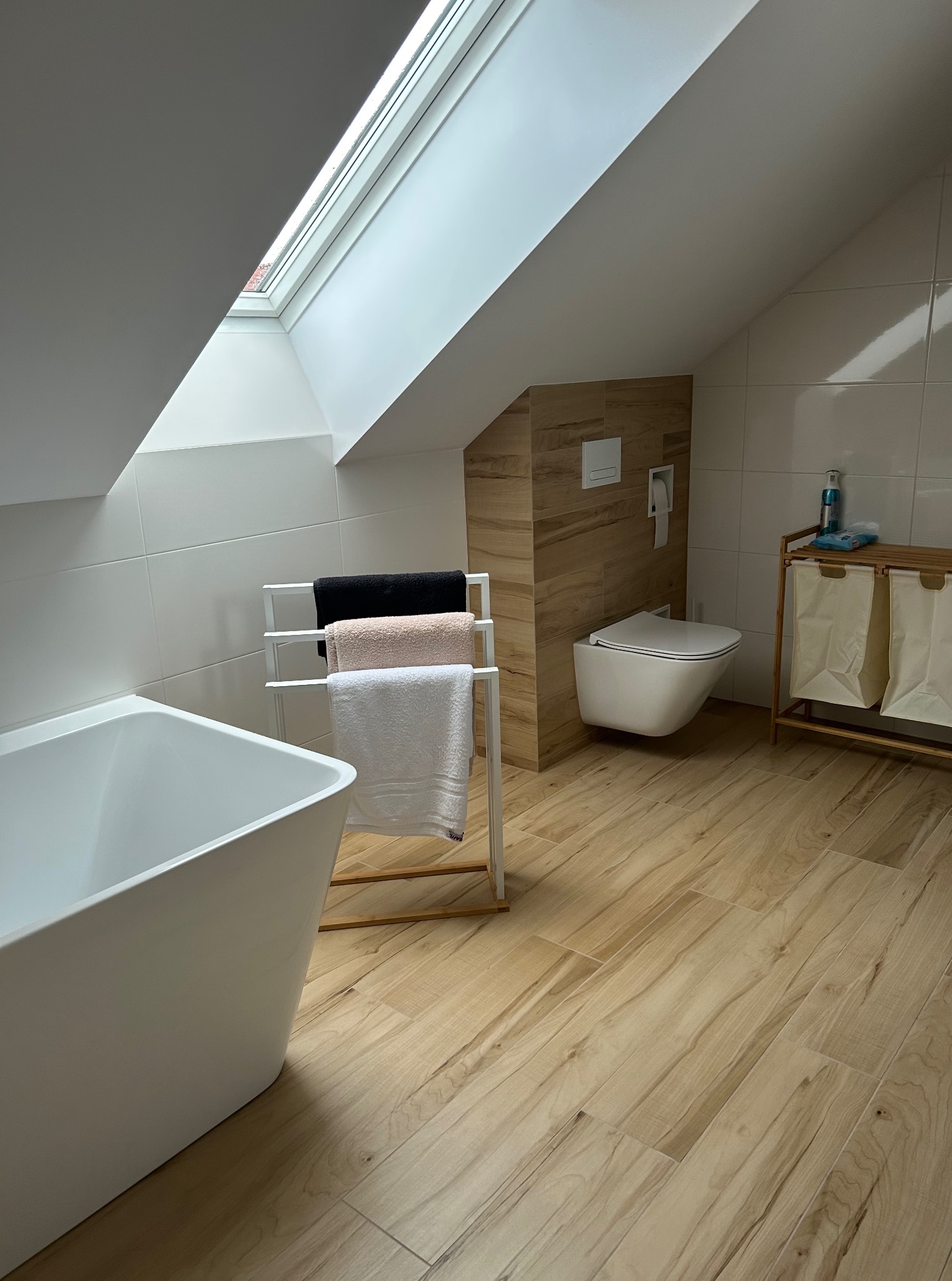 Jasna łazienka na poddaszu z drewnopodobną podłogą. Wolnostojąca wanna, podwieszana toaleta i okno dachowe tworzą nowoczesny, minimalistyczny wystrój.