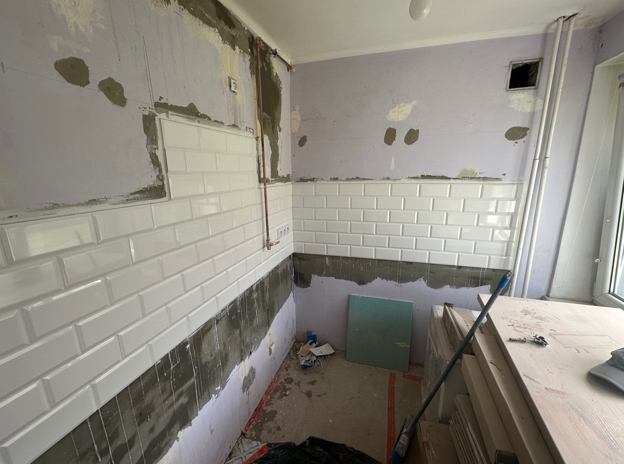 Białe, prostokątne płytki częściowo ułożone na ścianie w trakcie remontu kuchni. Widoczne przewody i przygotowane podłoże. Ściany w odcieniach fioletu.