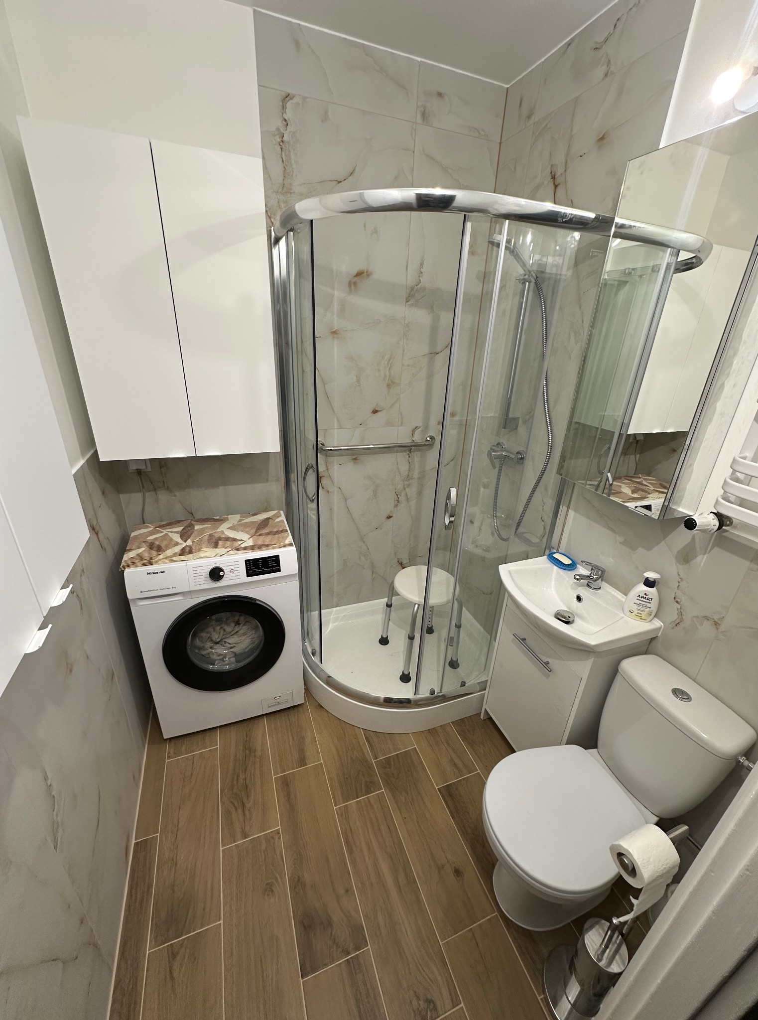 Funkcjonalna łazienka z kabiną prysznicową, pralką pod szafką i drewnopodobnymi płytkami na podłodze. Marmurowe płytki ścienne i biała armatura tworzą spójną całość.