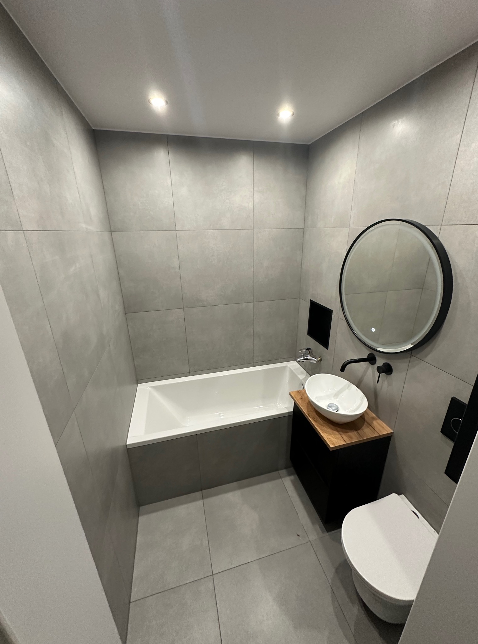 Szara łazienka z wanną, umywalką na szafce z drewnianym blatem, okrągłym lustrem i toaletą. Nowoczesny design, płytki imitujące beton.
